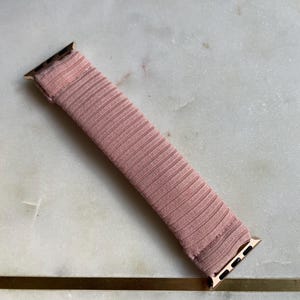 May include: A blush pink watch band with a ribbed texture. The band has gold-colored connectors on each end. The band is approximately 6 inches long.