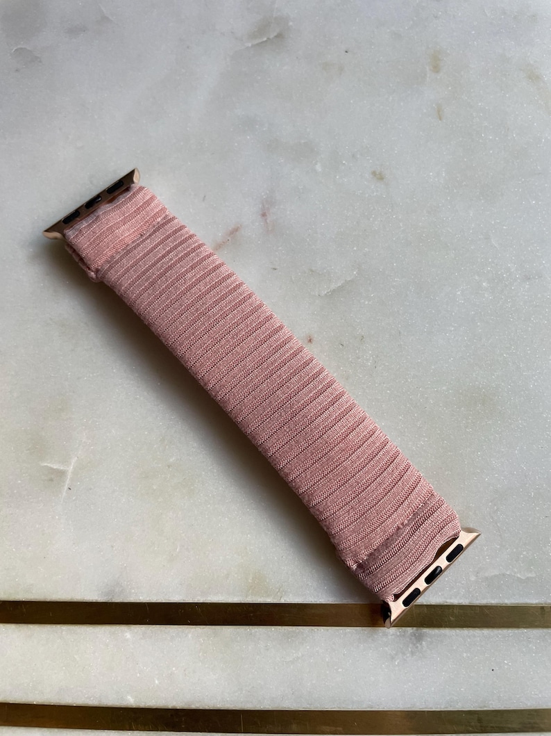 May include: A blush pink watch band with a ribbed texture. The band has gold-colored connectors on each end. The band is approximately 6 inches long.