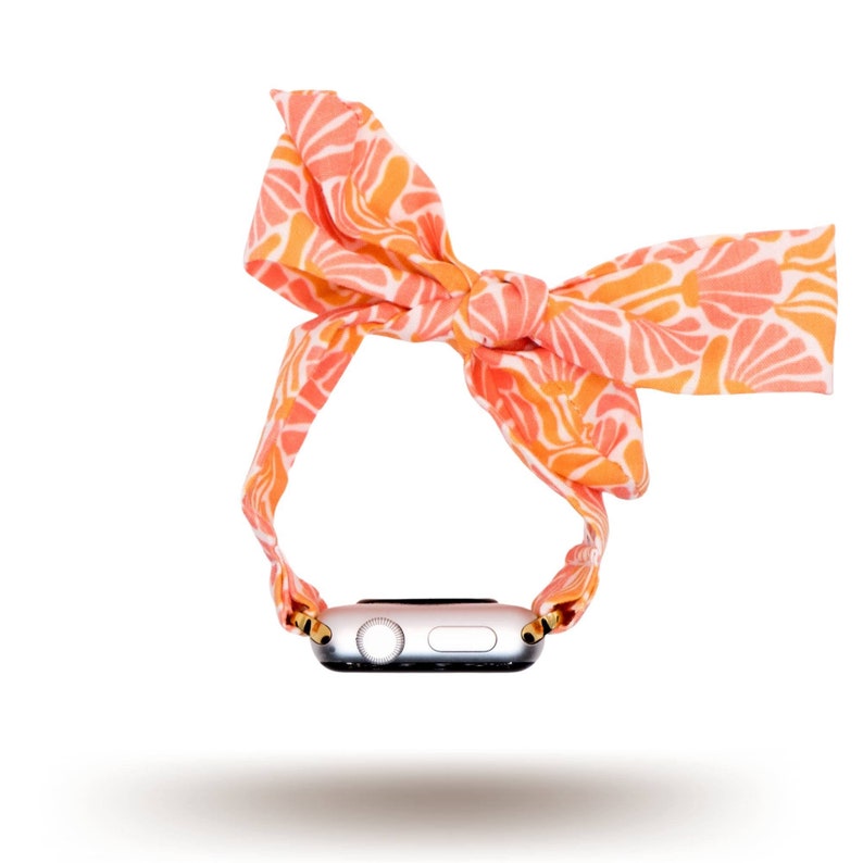 May include: A pink and orange patterned fabric watch band with a bow on top. The band is wrapped around a silver Apple Watch.