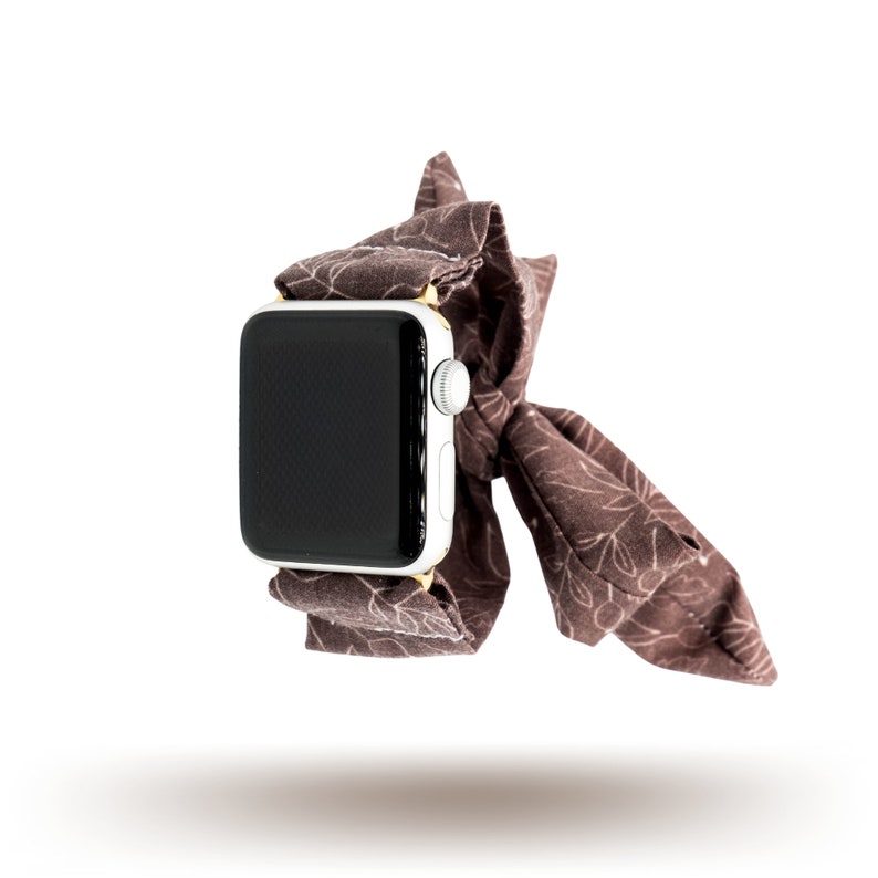 May include: A brown fabric watch strap with a white and gold Apple Watch. The strap is tied in a bow.
