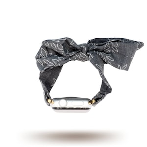 May include: A grey fabric Apple Watch band with a white leaf pattern and a gold buckle. The band is tied in a bow.