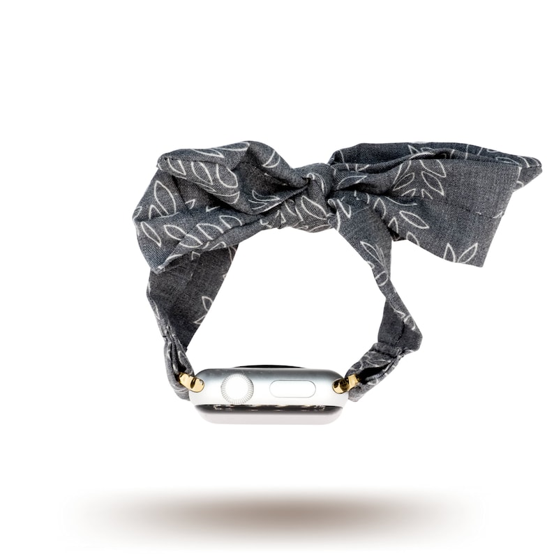 May include: A grey fabric Apple Watch band with a white leaf pattern and a gold buckle. The band is tied in a bow.
