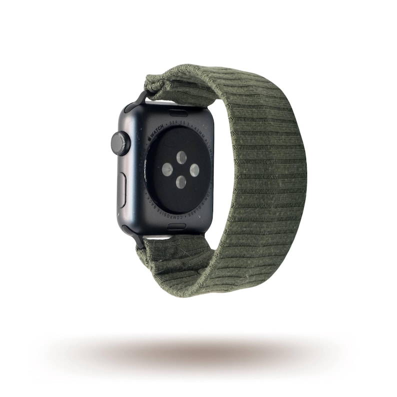 May include: A black smartwatch with a green ribbed fabric strap. The watch face is black with a circular button. The strap is a muted green colour and appears to be made of a soft, textured material.