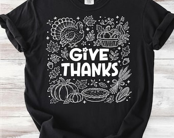 Może przedstawiać: Czarny t-shirt z krótkim rękawem z białym nadrukiem graficznym z napisem GIVE THANKS. Wzór zawiera ilustracje indyka, dyń, ciasta, jesiennych liści, żołędzi, kłosów pszenicy i kukurydzy, ułożone w kwadratowy układ.