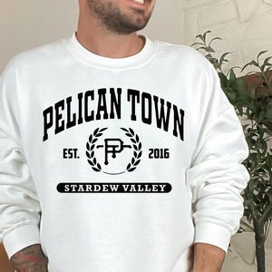 May include: White crewneck sweatshirt with the text "PELICAN TOWN" in a black, arched font. Below the text is "EST. 2016" and "STARDEW VALLEY". The design includes a laurel wreath and a stylized "P" in the center. The sweatshirt is a casual style.