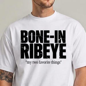 Op de afbeelding: Een wit T-shirt met korte mouwen voorzien van de zwarte tekst BONE-IN RIBEYE in een verweerd, gebarsten lettertype, met de kleinere zwarte tekst "my two favorite things" direct onder de hoofdafbeelding op de borst gedrukt.