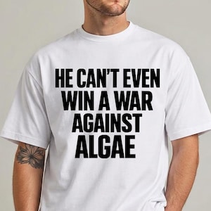 Könnte beinhalten: Ein weißes T-Shirt mit kurzen Ärmeln und einem fetten schwarzen Text auf der Brust, der lautet: HE CAN'T EVEN WIN A WAR AGAINST ALGAE. Das Shirt hat einen Rundhalsausschnitt und eine Standardpassform für einen schlichten Look.