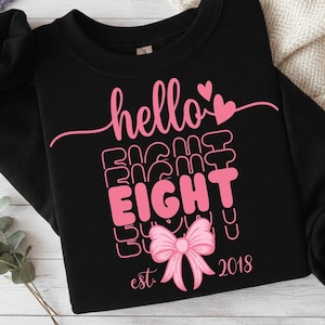 Op de afbeelding: Zwart sweatshirt met roze tekst "hello EIGHT", een strik-afbeelding en het jaar "2018". Het ontwerp bevat harten en het woord "est.". Het sweatshirt is op een licht oppervlak gevouwen.