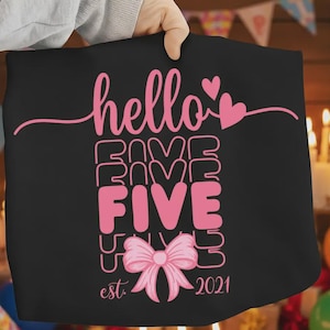 Puede incluir: Camiseta negra con texto rosa "hello FIVE" en un diseño apilado. También se presenta un lazo rosa y el año "2021". La camiseta está sujeta por una persona.