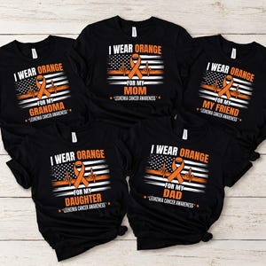 May include: Five black t-shirts featuring a graphic with an orange awareness ribbon, a heartbeat line, and a stylized American flag. Each shirt displays the text I WEAR ORANGE FOR MY followed by a family member or friend and LEUKEMIA CANCER AWARENESS.