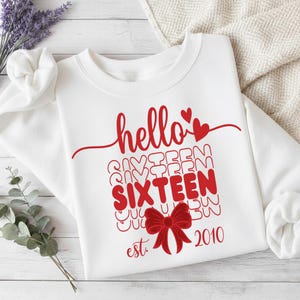 May include: A white crewneck sweatshirt featuring red graphic text that reads hello SIXTEEN est. 2010. The design includes a red bow illustration, two small red hearts, and a repeating wavy line pattern behind the word SIXTEEN in red ink.