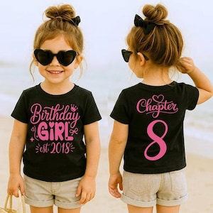 May include: A black t-shirt with pink text that says "Birthday Girl est 2018" on the front and "Chapter 8" on the back. The girl is wearing heart-shaped sunglasses and khaki shorts. The image is taken outdoors.