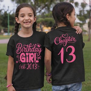 May include: Two girls wearing black t-shirts with pink text. One shirt says "Birthday GIRL est. 2013" and the other says "Chapter 13". Both girls are wearing blue jeans and bracelets. The shirts are for a birthday.