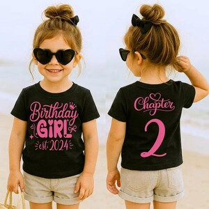 May include: Black t-shirt with pink text that reads "Birthday Girl est 2024" on the front and "Chapter 2" on the back. The shirt is worn by a child wearing heart-shaped sunglasses and khaki shorts.