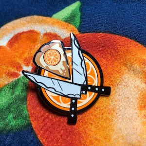 May include: A black and white enamel pin featuring two knives crossing over a slice of orange pie. The pin is on a blue and orange fabric background.
