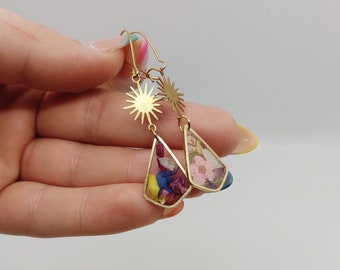 May include: Gold earrings with a sunburst design and teardrop-shaped pendants filled with pressed flowers. The flowers are in shades of pink, blue, yellow, and purple.