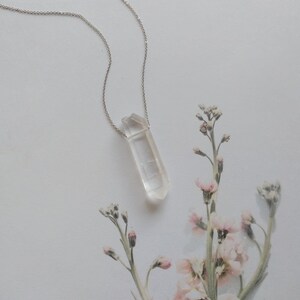 May include: A silver chain necklace with a clear quartz crystal pendant. The pendant is a long, thin, pointed crystal.