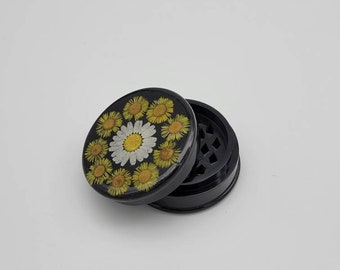 Può includere: Macinino nero con un coperchio rotondo nero con un motivo floreale di margherite gialle e bianche.