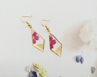 May include: A pair of gold earrings with a diamond-shaped design. Each earring features a pressed pink flower encased in clear resin.