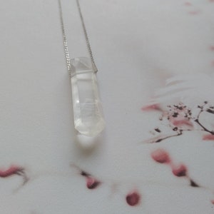 May include: A clear quartz crystal pendant on a silver chain. The crystal is faceted and has a pointed tip. The pendant is hanging against a white background with pink floral patterns.