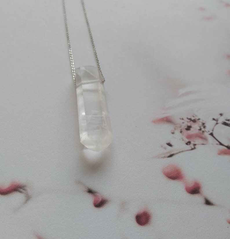 May include: A clear quartz crystal pendant on a silver chain. The crystal is faceted and has a pointed tip. The pendant is hanging against a white background with pink floral patterns.