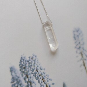 May include: A clear quartz crystal pendant on a silver chain. The pendant is a long, thin, pointed shape.
