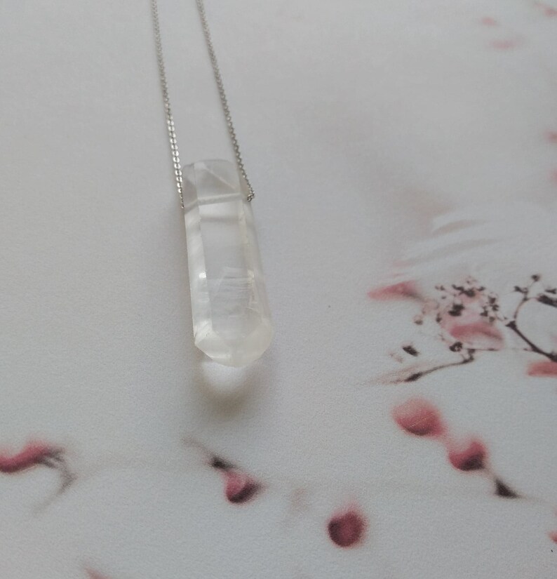 May include: A clear quartz crystal pendant on a silver chain. The crystal is faceted and has a pointed tip. The pendant is hanging against a white background with pink floral patterns.