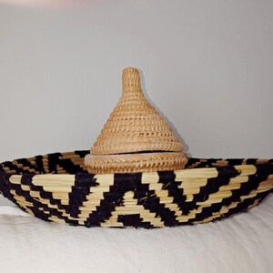 May include: A woven basket with a black and natural geometric pattern. A smaller, lidded basket sits inside. The lid is cone-shaped. The baskets are made of natural fibres and are likely handmade.