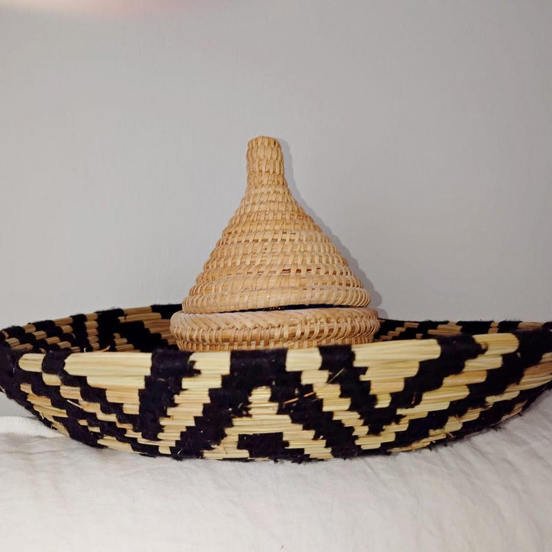 May include: A woven basket with a black and natural geometric pattern. A smaller, lidded basket sits inside. The lid is cone-shaped. The baskets are made of natural fibres and are likely handmade.