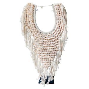Puede incluir: Un collar de conchas blancas con flecos de cuerda blanca. El collar se muestra en un soporte negro.