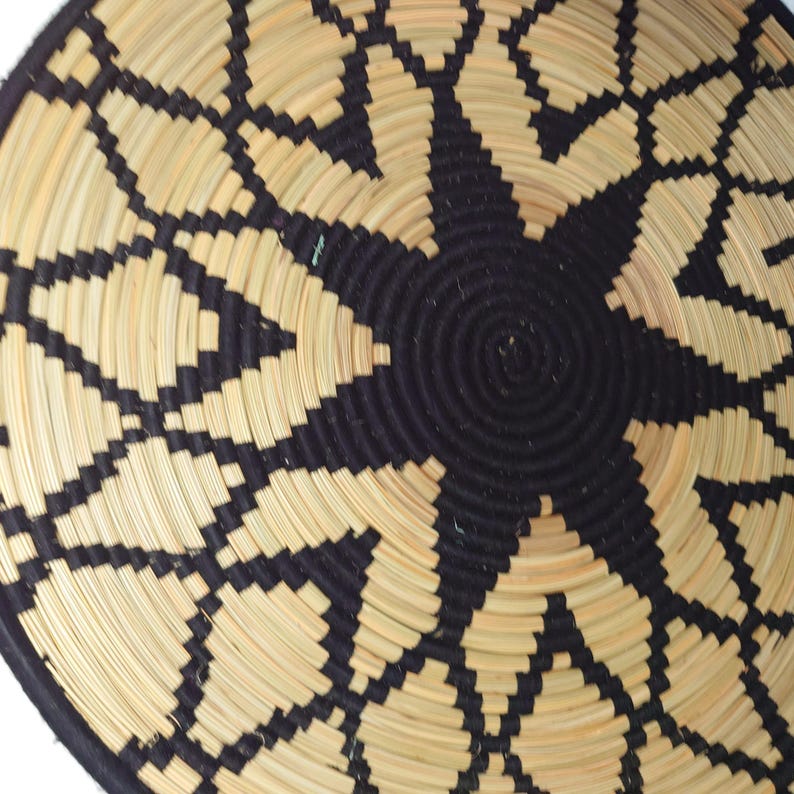May include: A woven basket with a starburst design. The basket features a central black spiral surrounded by a pattern of black and tan geometric shapes. The rim of the basket is black.