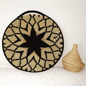May include: A large, circular woven basket featuring a black starburst pattern and a black handle. Beside it is a small, conical woven basket with a lid. Both baskets are crafted from natural and black fibres.
