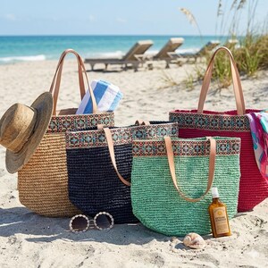 K&ouml;nnte beinhalten: Vier gewebte Strandtaschen in Natur, Marineblau, Gr&uuml;n und Rot. Jede Tasche hat ein dekoratives Band mit geometrischem Muster und Ledergriffen. Ein Strohhut, Sonnenbrille, Sonnencreme und eine Muschel liegen im Sand.