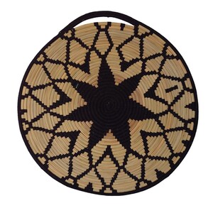 May include: Circular woven basket featuring a black and natural geometric starburst pattern. The basket has a black handle and rim. The natural colour is a light tan. The basket is made of woven natural fibres.