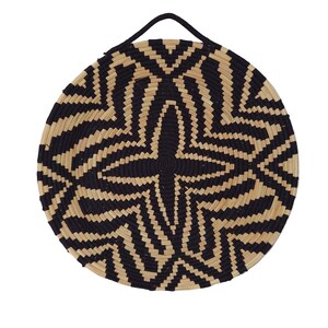 May include: Circular woven basket with a black and beige geometric design. The basket has a black handle at the top for hanging. The pattern features a starburst in the centre, with radiating lines. The basket is made of natural fibres.