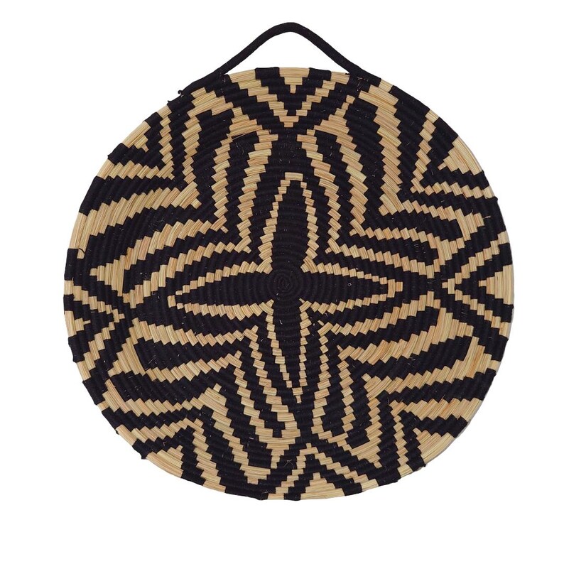 May include: Circular woven basket with a black and beige geometric design. The basket has a black handle at the top for hanging. The pattern features a starburst in the centre, with radiating lines. The basket is made of natural fibres.