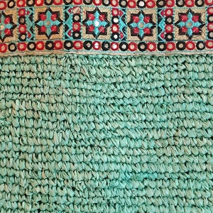 K&ouml;nnte beinhalten: Nahaufnahme eines gewebten Textils mit t&uuml;rkisfarbenem Hintergrund. Der obere Rand zeigt ein besticktes Band mit geometrischen Mustern in Rot, Schwarz, T&uuml;rkis und Gold, mit kleinen Spiegelakzenten. Der untere Bereich hat ein strukturiertes, gewebtes Aussehen.