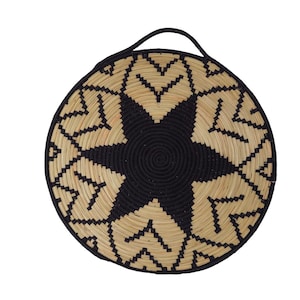 May include: Circular woven basket featuring a black starburst pattern and a black border. The basket has a natural tan hue with black detailing and a black handle. Ideal for wall decoration, this basket is a stylish addition to any home.