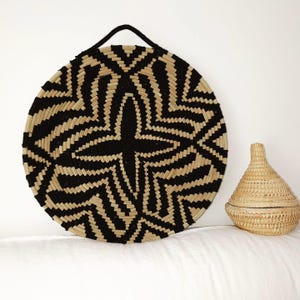 May include: A circular woven basket with a black and natural geometric design and a black handle. A smaller, lidded basket is next to it. The baskets are made of natural fibres and are on a white surface.
