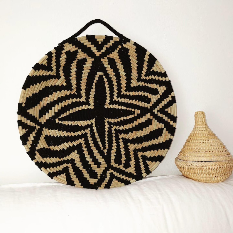 May include: A circular woven basket with a black and natural geometric design and a black handle. A smaller, lidded basket is next to it. The baskets are made of natural fibres and are on a white surface.