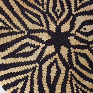 May include: A round woven basket with a geometric design. The pattern consists of alternating black and natural beige bands, emanating from a central spiral. The basket's texture and colour scheme provide a strong visual contrast.