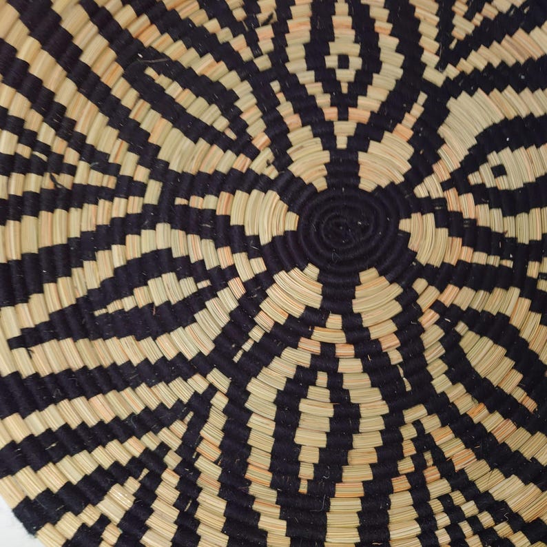 May include: A round woven basket with a geometric design. The pattern consists of alternating black and natural beige bands, emanating from a central spiral. The basket's texture and colour scheme provide a strong visual contrast.