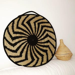 May include: A large, circular woven basket with a black and natural zig-zag design and a black handle. Beside it is a small, lidded basket with a conical shape, also woven from natural fibres. Ideal for home decor.