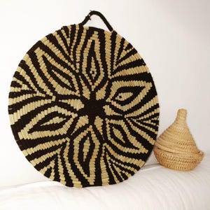 May include: A large, round woven wall basket with a black and natural fibre geometric design. A smaller, lidded basket sits to the right. The baskets are made of natural fibres and are suitable for home decor.