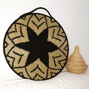 May include: A large, circular woven basket featuring a black starburst pattern and a black handle. Beside it is a small, lidded basket with a conical form. Both baskets are crafted from natural and black woven materials.