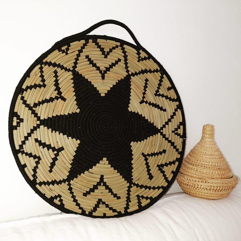 May include: A large, circular woven basket featuring a black starburst pattern and a black handle. Beside it is a small, lidded basket with a conical form. Both baskets are crafted from natural and black woven materials.