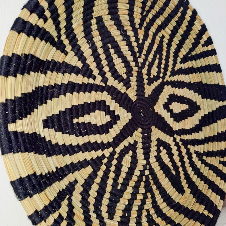 May include: A circular woven basket with a bold geometric design. It features alternating bands of black and natural beige fibres, forming a radial pattern. The centre has a tightly coiled, dark black circle.