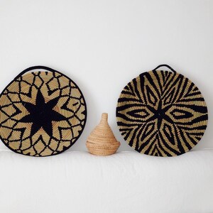 May include: Three woven decorative items. Two large, round wall hangings with black and beige geometric patterns. A small, lidded basket sits between them. The items are made of natural fibres and have a rustic aesthetic.