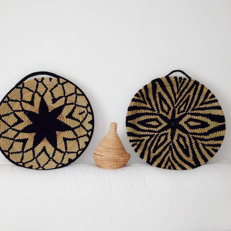 May include: Three woven decorative items. Two large, round wall hangings with black and beige geometric patterns. A small, lidded basket sits between them. The items are made of natural fibres and have a rustic aesthetic.