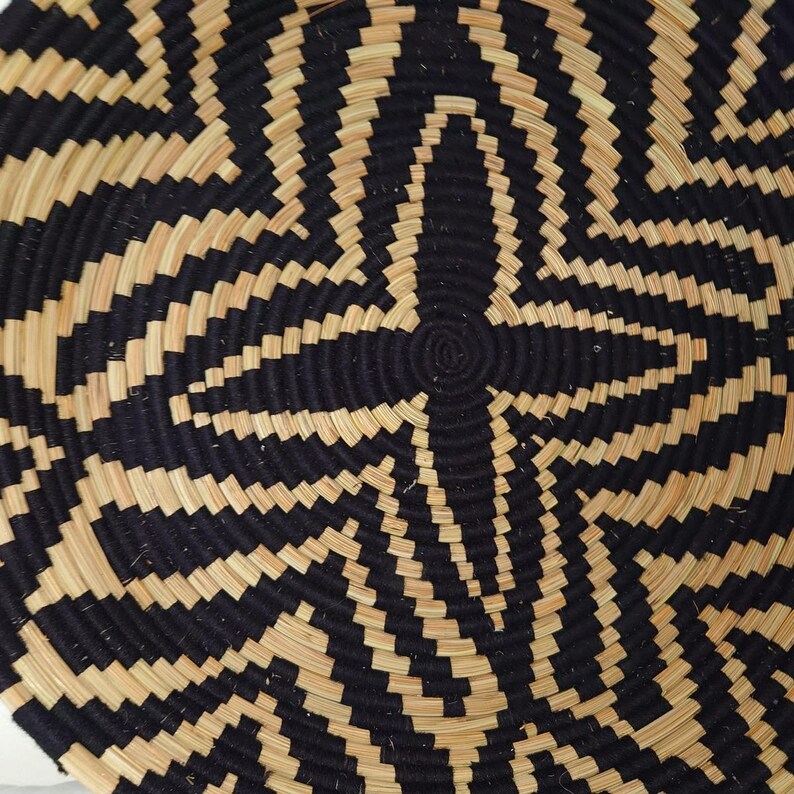 May include: Close-up of a round woven basket with a geometric pattern. The basket features a repeating design of black and natural-coloured woven material, creating a starburst effect. The basket is likely made of natural fibres.